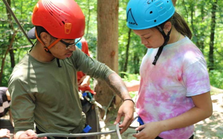 An outward bound instructor helps a student tie a rope to their harness. They are both wearing helmets and stand in a wooded area. 